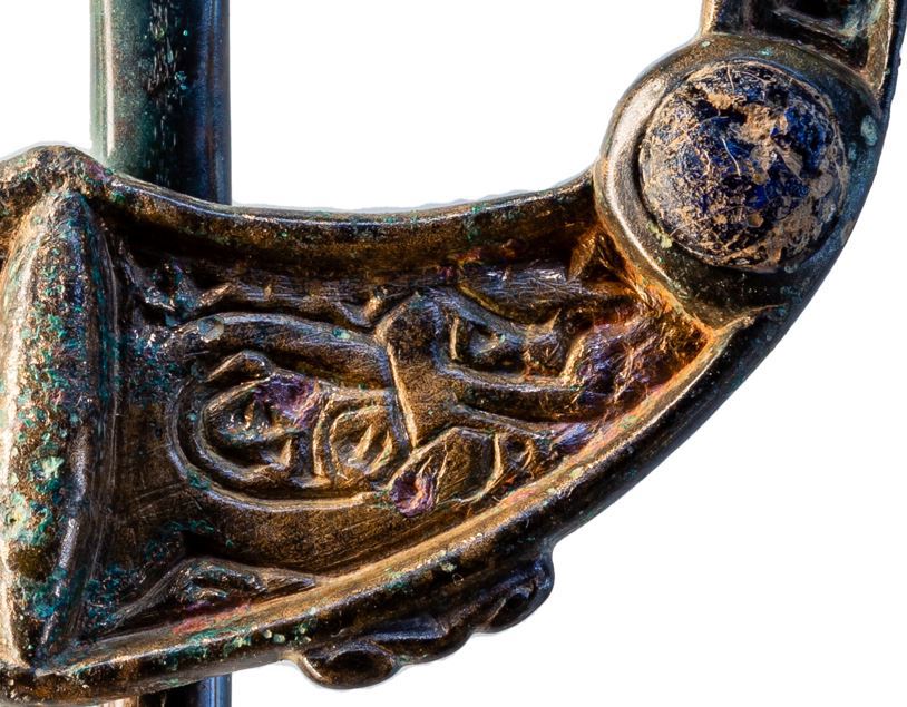 Illustration 12: Close-up of the left terminus showing the recessed panel and raised zoomorphic design bordered at the ‘terminus’ end by a raised lentoid shape and a blue-glass stud at the hoop end, with a bronze flourish along the outside. The red-coloured markings are a result of scarring from cuprous oxide blisters. (Photo: John Channing, AMS).